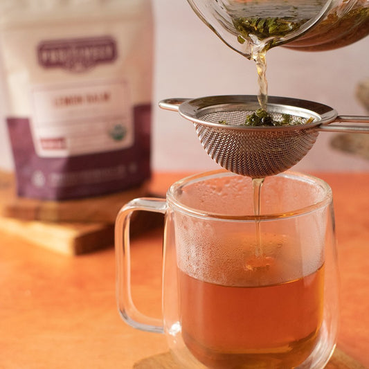 Positively Botanicals Organic Lemon Balm tea being poured through a strainer into a glass mug, highlighting its soothing, caffeine-free infusion.