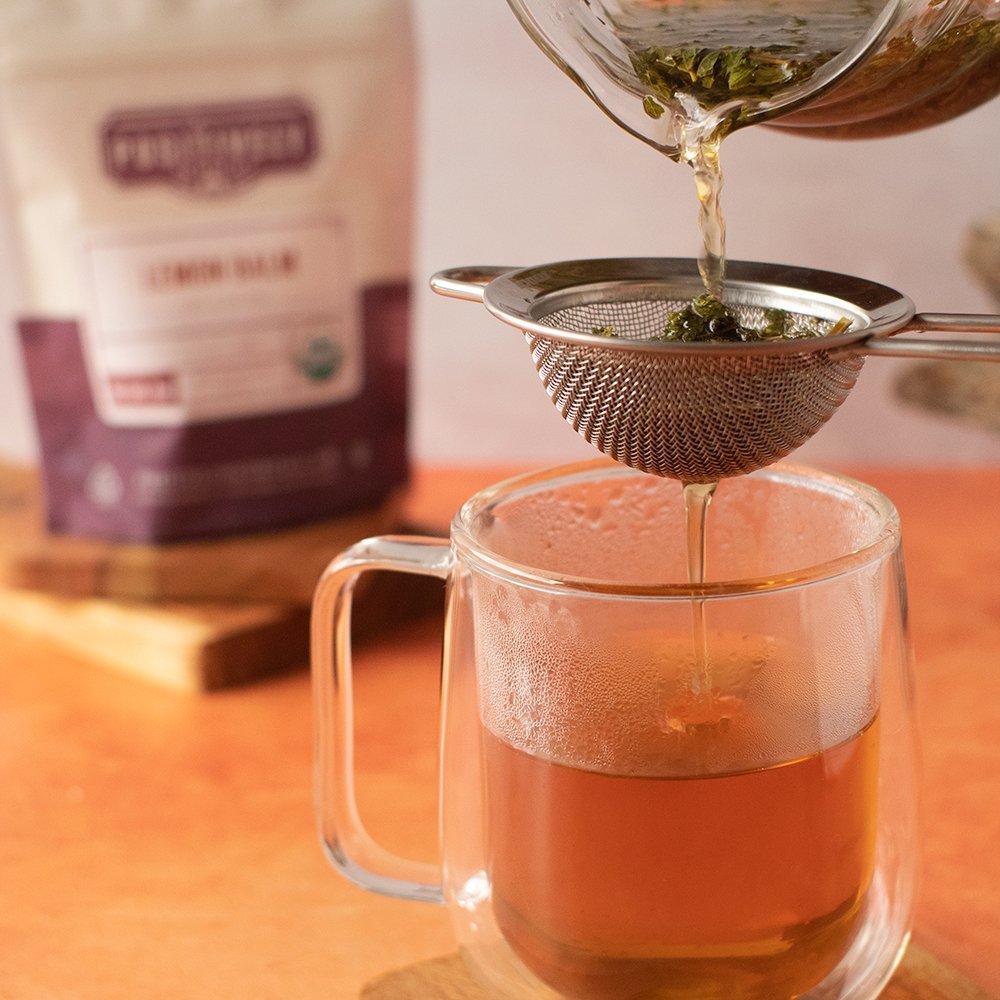 Positively Botanicals Organic Lemon Balm tea being poured through a strainer into a glass mug, highlighting its soothing, caffeine-free infusion.