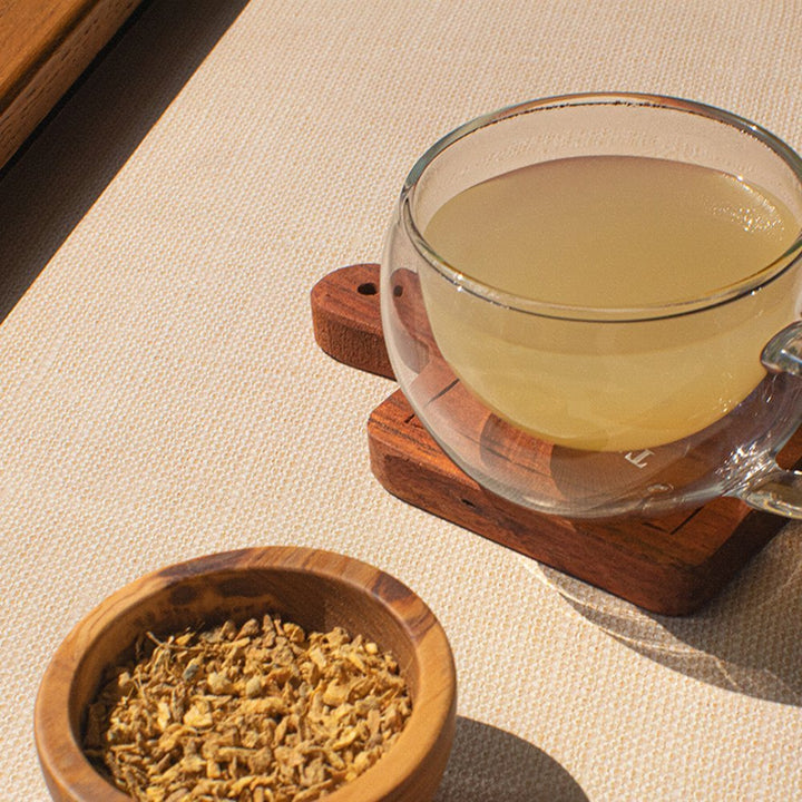 A glass cup of brewed ginger root tea placed on a wooden coaster next to a small wooden bowl of dried ginger root pieces on a beige surface.
