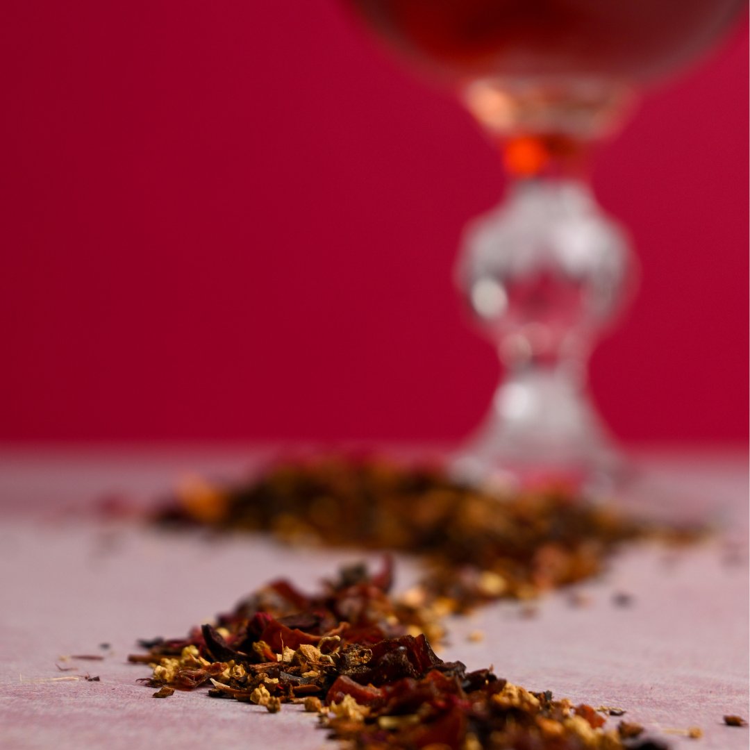 Raspberry Rosé organic herbal tea leaves artfully arranged with a glass of steeped tea blurred in the background.