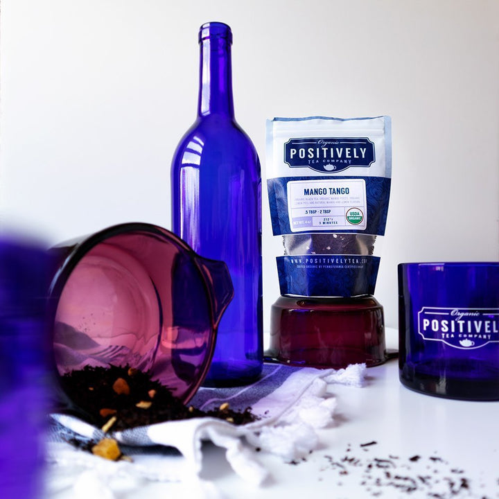 Positively Tea Company Mango Tango black tea packaging displayed with scattered tea leaves and vibrant glassware, evoking a bold tropical vibe.